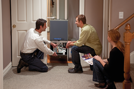 An HVAC technician explaining a furnace system to homeowners, showing the open unit and its components during a consultation inside their home.