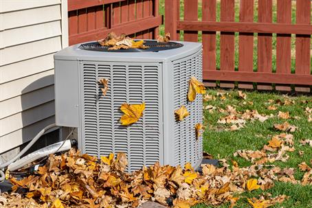 Outdoor air conditioner in a backyard with autumn leaves on and around it, illustrating the need for fall HVAC maintenance.