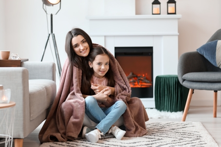 A mother and daughter wrapped in a cozy blanket sitting in front of a glowing fireplace, symbolizing warmth, comfort, and family time during cold weather.